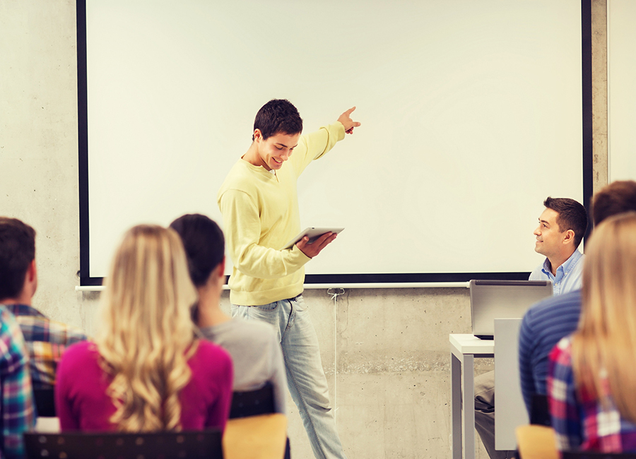 A student confidently presenting in a classroom while pointing at a blank screen, engaging an audience of classmates and a teacher.