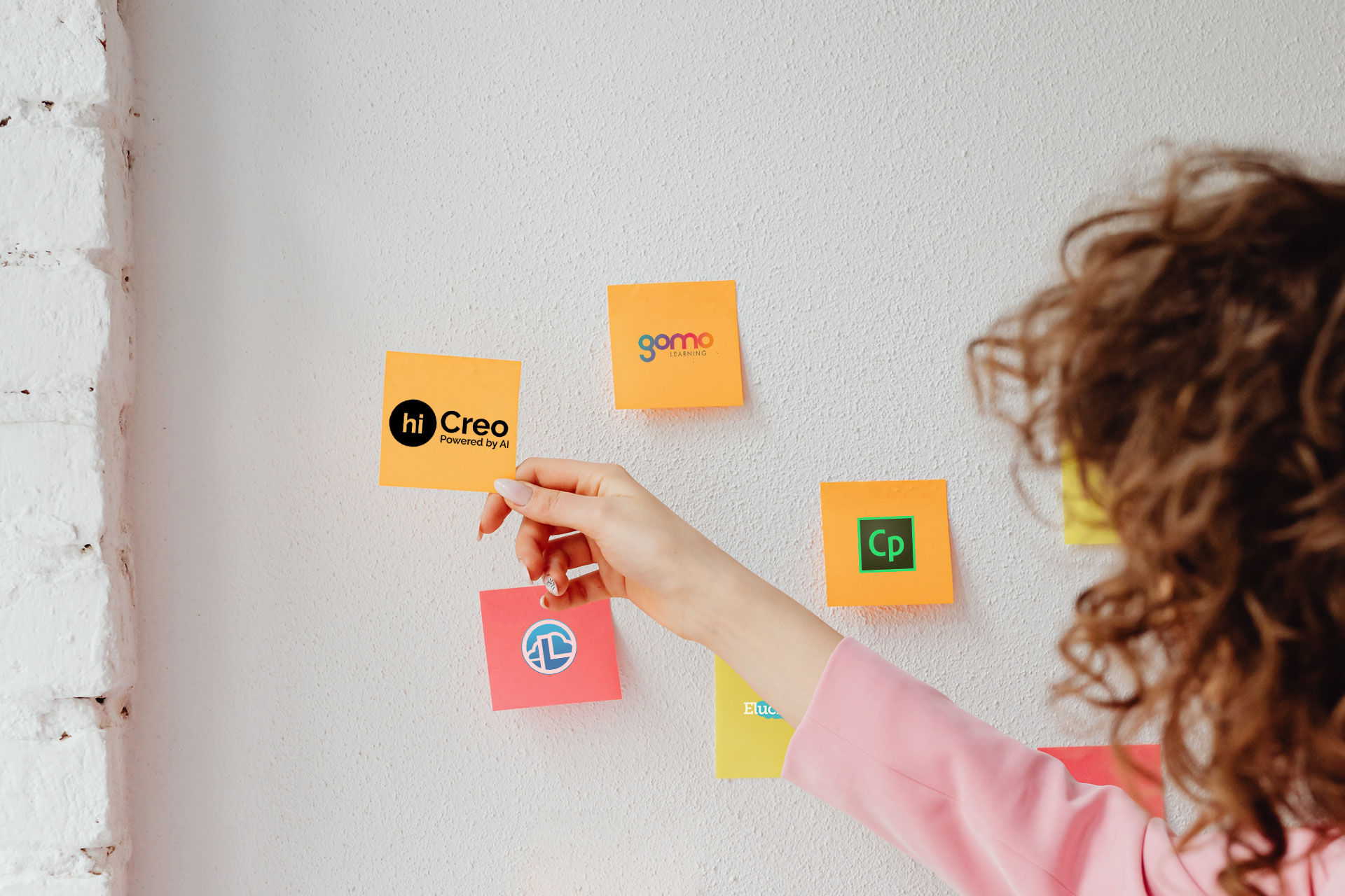 A woman is selecting a sticky note with the hiCreo logo from a wall covered with sticky notes. The other sticky notes on the wall have the logos of Captivate, Gomo, and Lectora, suggesting a comparison of the top 5 eLearning tools.